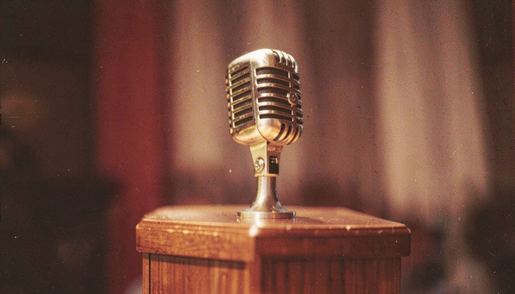 A close-up film photograph of a vintage microphone on a wooden podium, warm brown and gold tones, shallow depth of field with soft grain