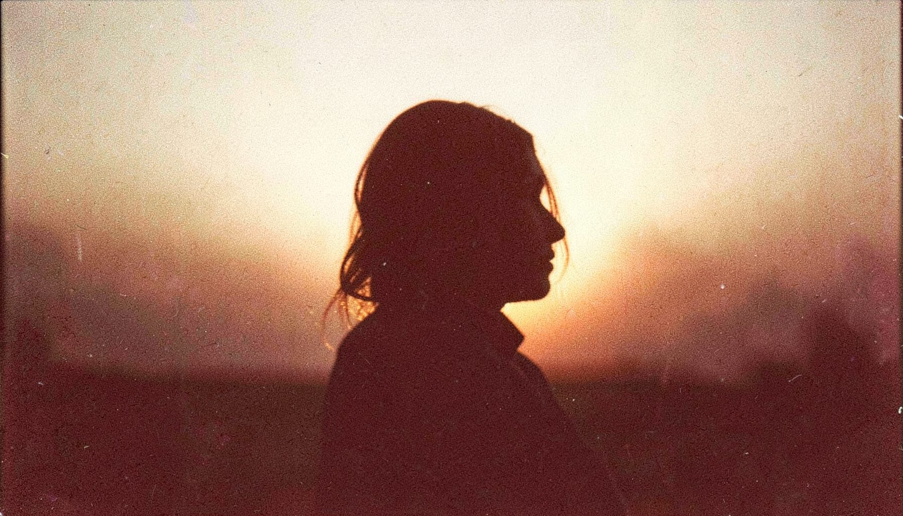 A grainy film photograph of a woman's silhouette against a warm golden sunset, soft earth tones and amber light