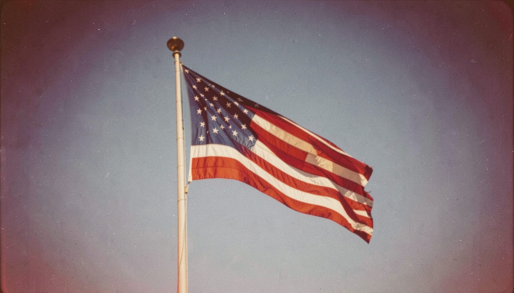 A warm, softly lit photograph of an American flag waving against a clear blue sky, with rich red and navy tones and visible film grain.