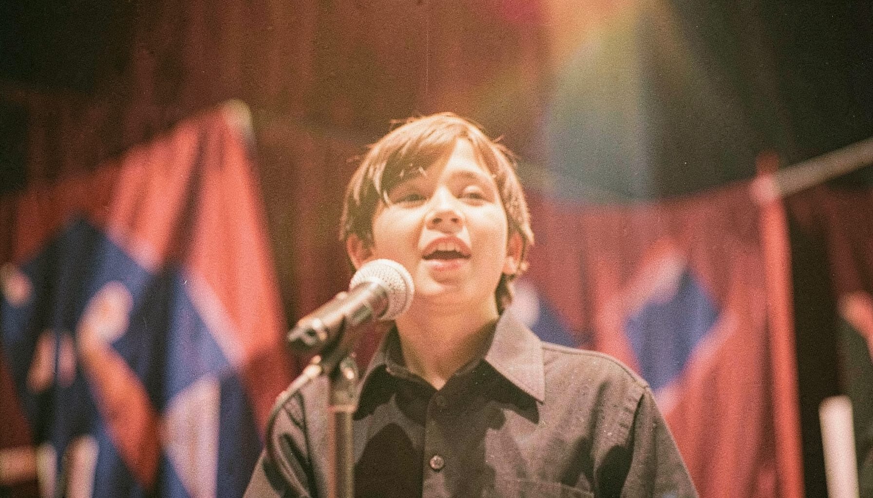 A close-up analog photograph of a young person speaking at a microphone on a large convention stage, red and blue bunting in the background, warm amber stage lighting, visible film grain.