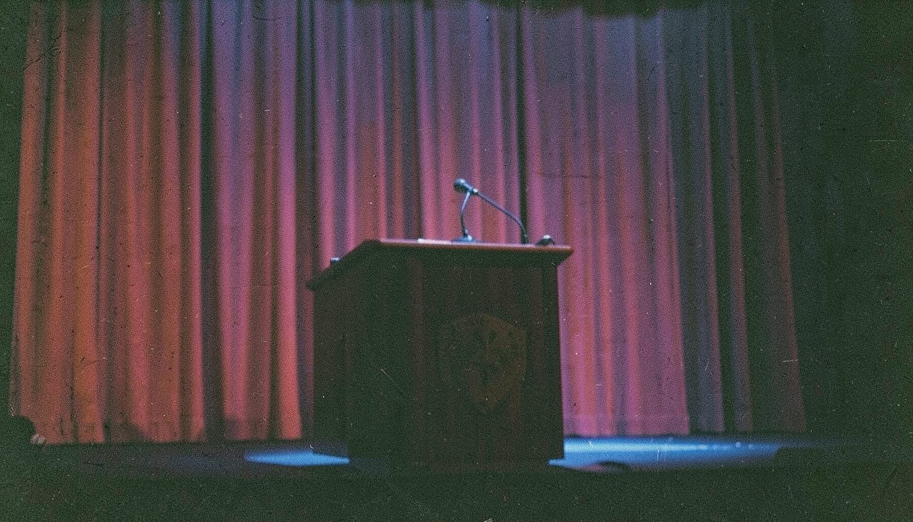 A grainy film photograph of a podium with microphones on a convention stage, deep red curtain behind, blue stage lighting casting dramatic shadows.