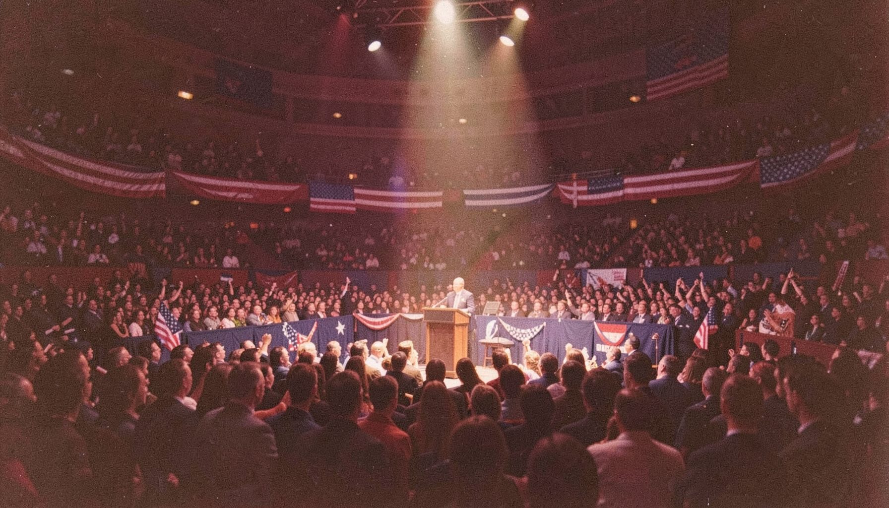 A wide banner photograph of a packed political convention hall, deep red and navy blue tones, American flags draped across the stage, warm spotlight glow on the podium, grainy film texture.