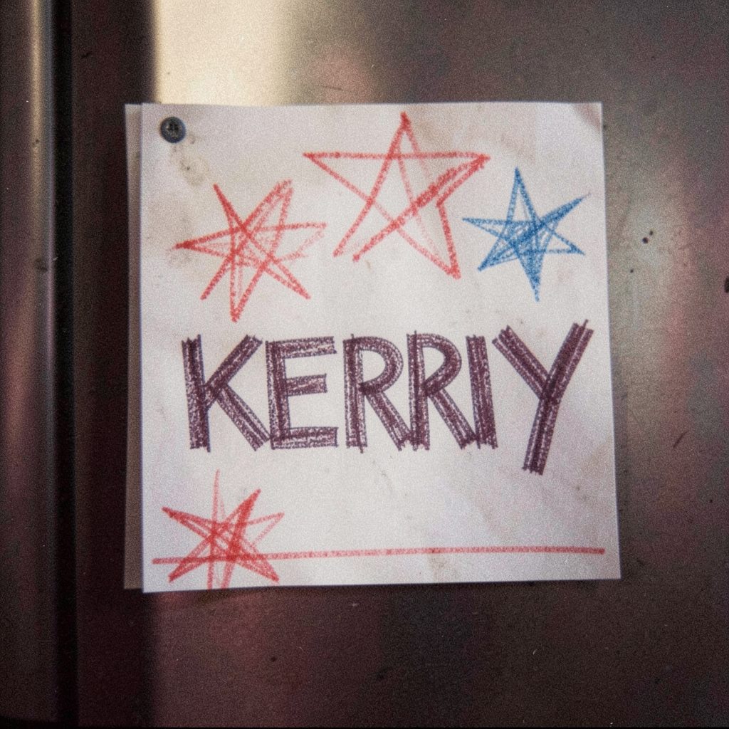 A grainy close-up of a child's crayon drawing on construction paper, showing red and blue stars and the word KERRY in bold block letters, taped to a refrigerator door.