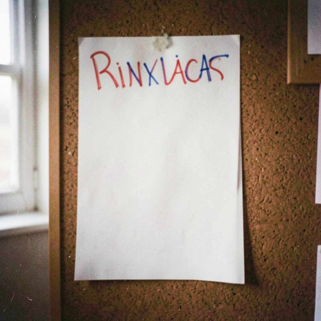 A grainy analog photograph of a hand-drawn campaign poster taped to a corkboard, featuring red and blue marker lettering against white poster board, with soft natural light from a nearby window.