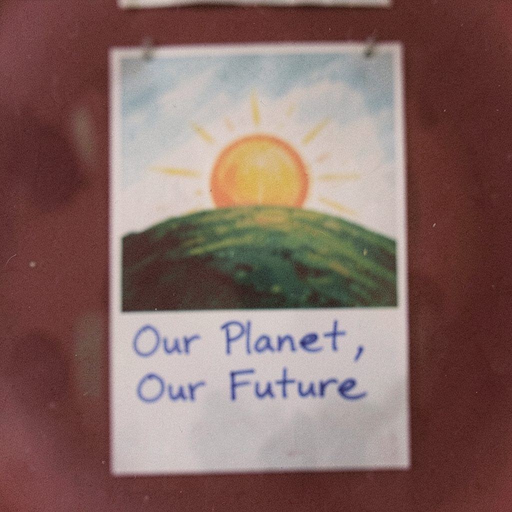 A soft-focus film photograph of a campaign poster with a painted sun rising over a green hill, the words 'Our Planet, Our Future' written in blue marker below.
