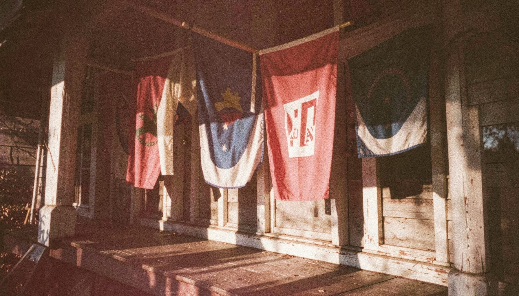 A warm, grainy photograph of red, white, and blue campaign banners hanging from a sunlit porch, with soft afternoon light casting long shadows across weathered wood.