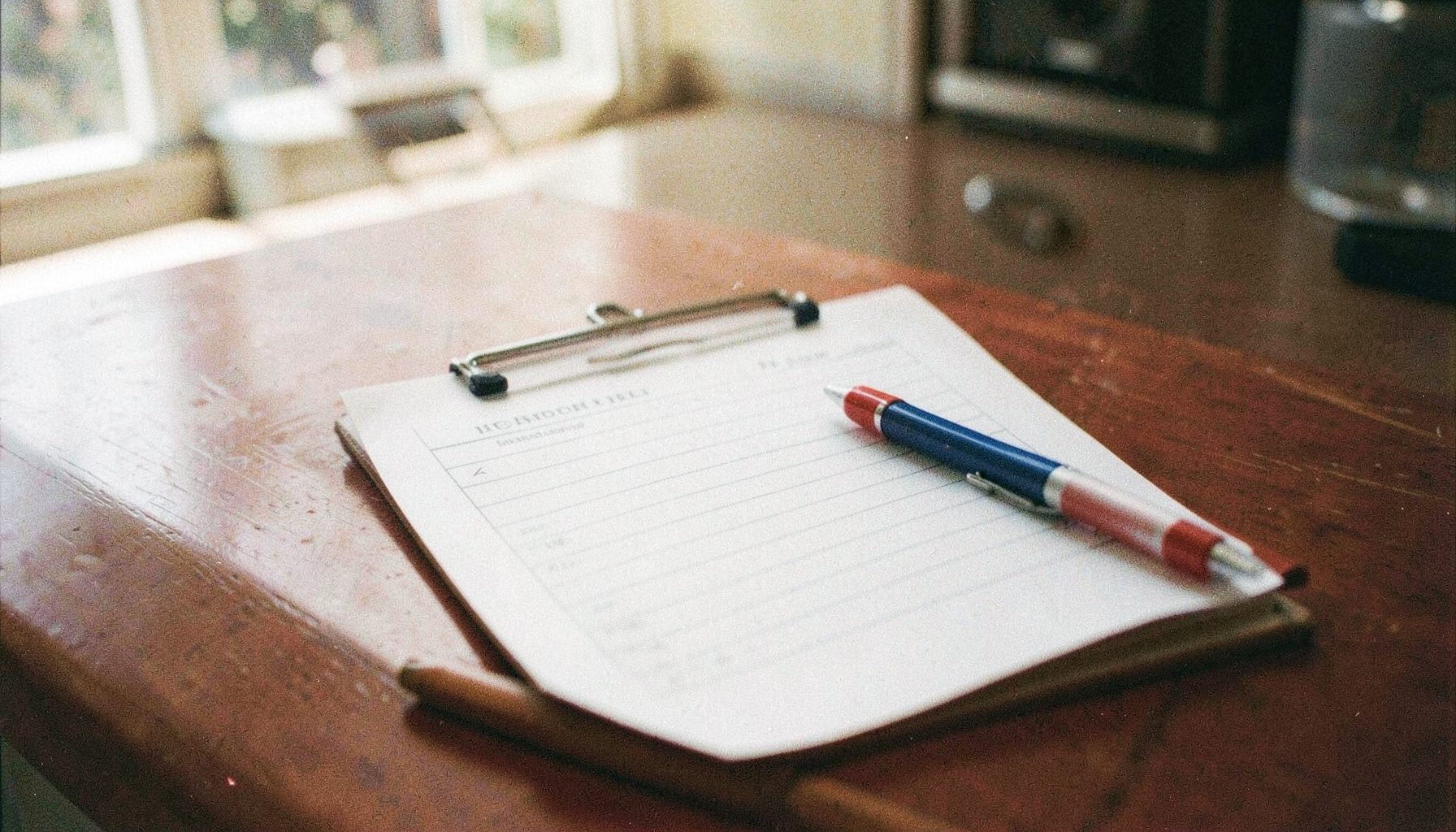 A grainy film photograph of a clipboard with a sign-up sheet and a red, white, and blue pen resting on top, sitting on a sunlit kitchen table.