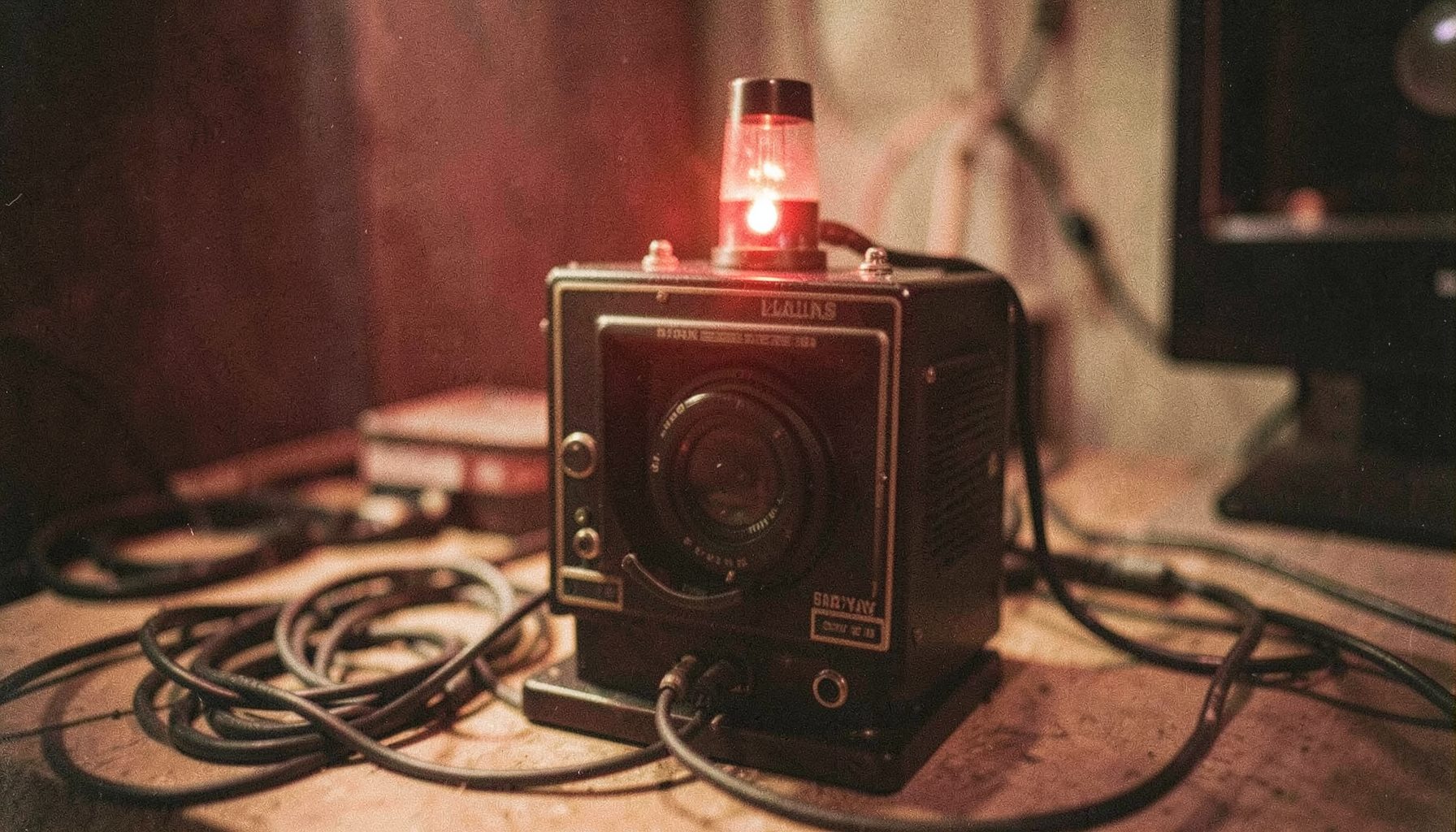 A close-up analog photograph of a vintage television studio camera with a red tally light glowing, cables coiled on the floor, warm tungsten lighting, shallow depth of field, visible film grain