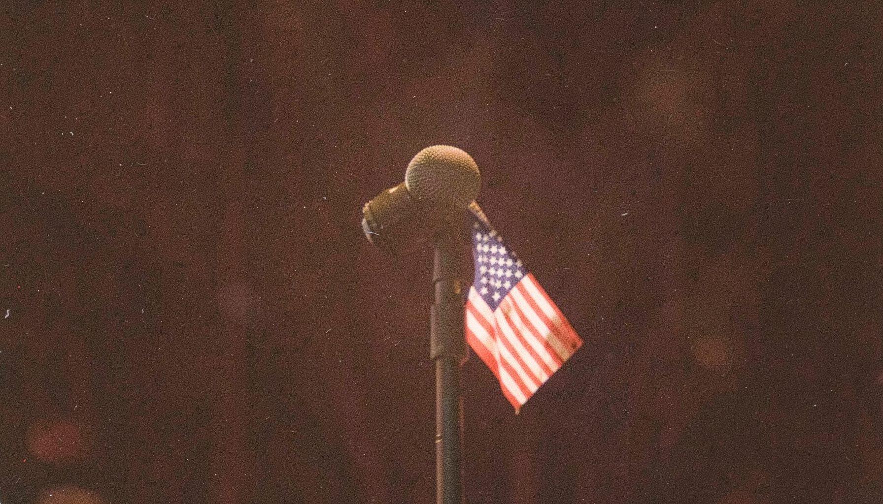 A grainy film photograph of a handheld microphone with a small American flag clipped to the stand, soft amber stage light, dark background