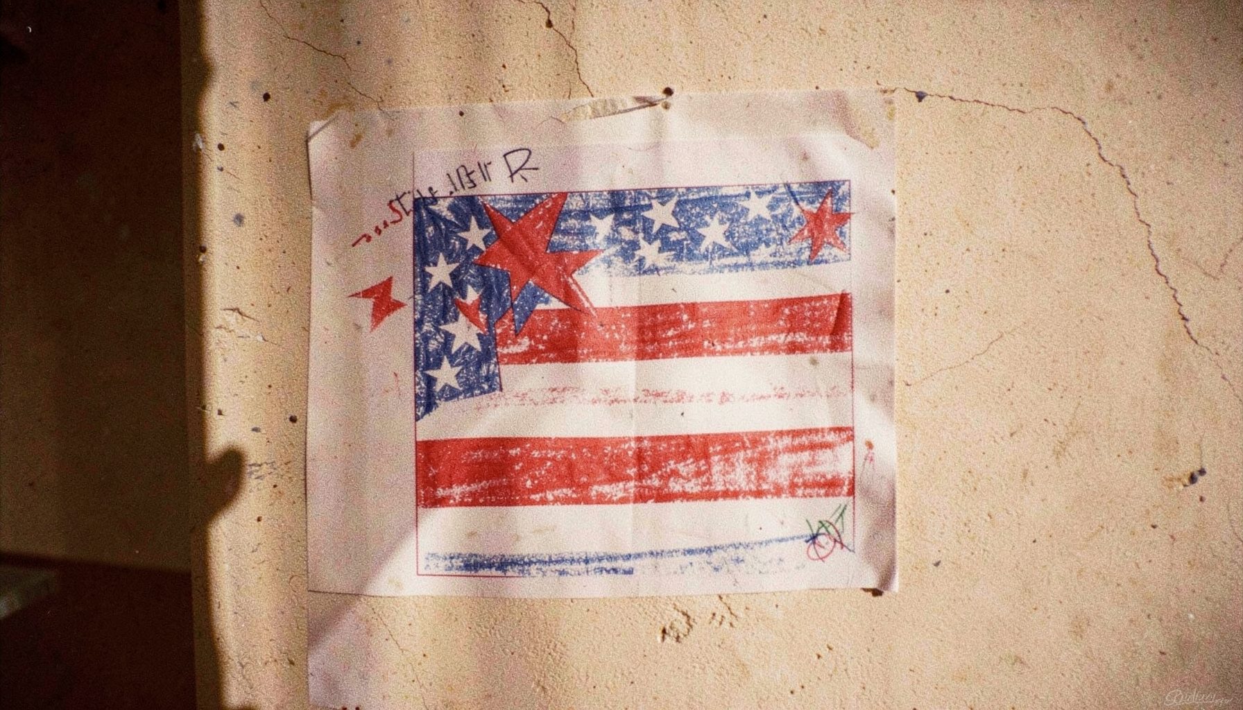 A film photograph of a child's hand-drawn campaign poster with stars and stripes in red, white, and blue, taped to a sunlit wall with visible grain.