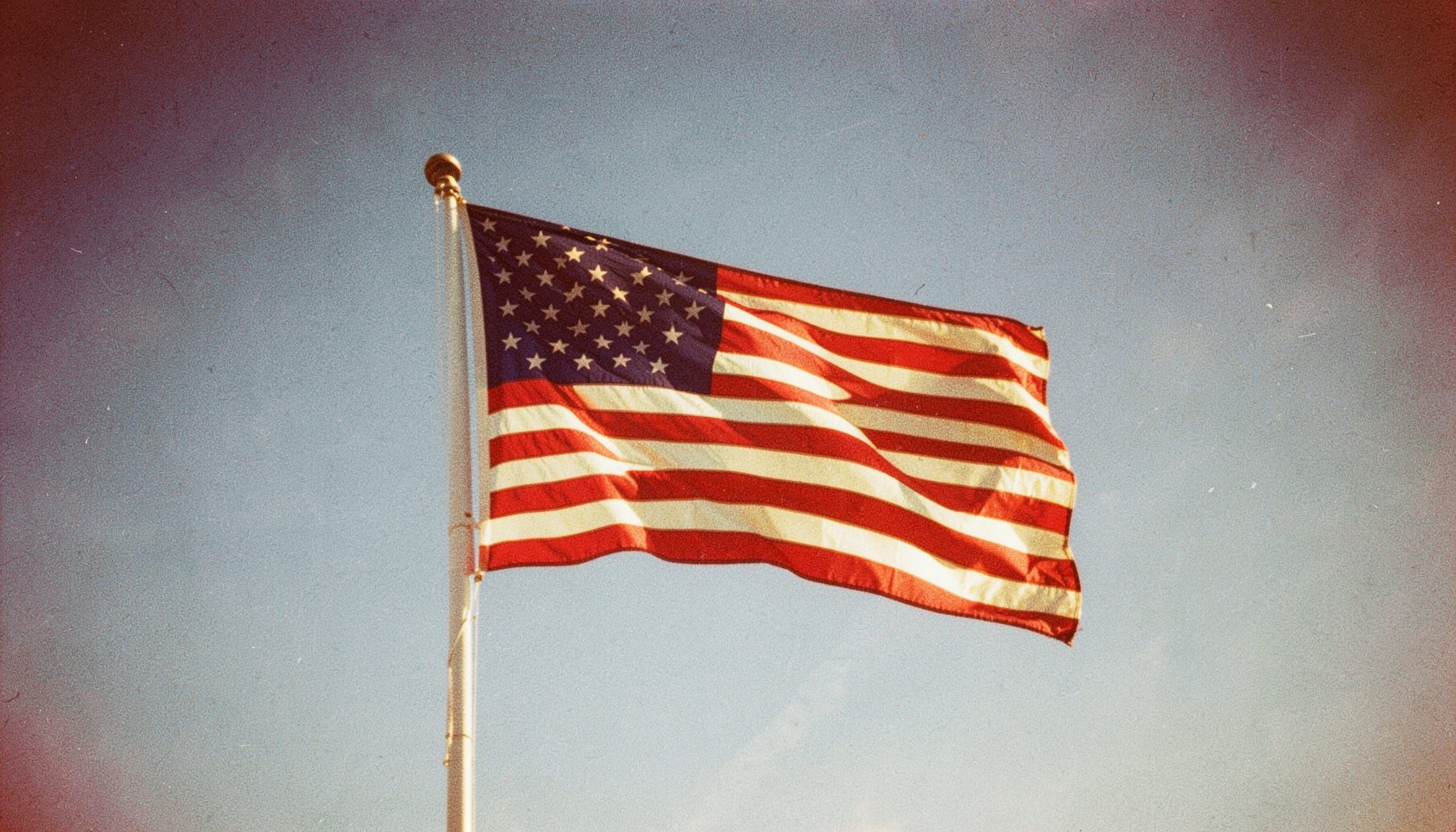 A wide photograph of a sunlit American flag waving against a bright blue sky, with warm golden and red tones and visible film grain.