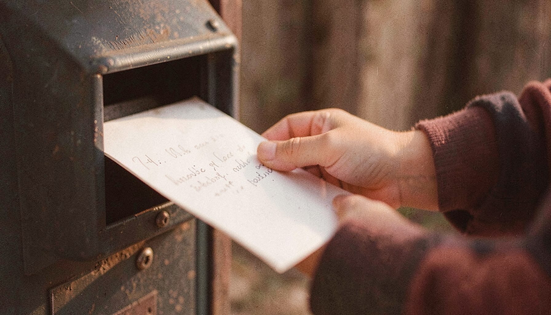 A warm-toned analog photograph of a child's hand placing a handwritten letter into a mailbox, soft afternoon light, visible film grain.