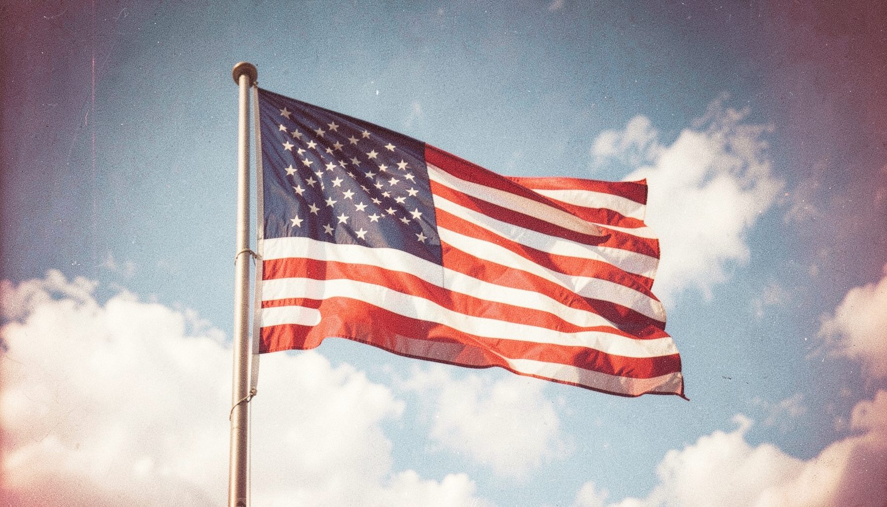 A wide banner photograph of a sunlit American flag waving against a bright blue sky with soft white clouds, warm and hopeful in tone, with visible film grain.