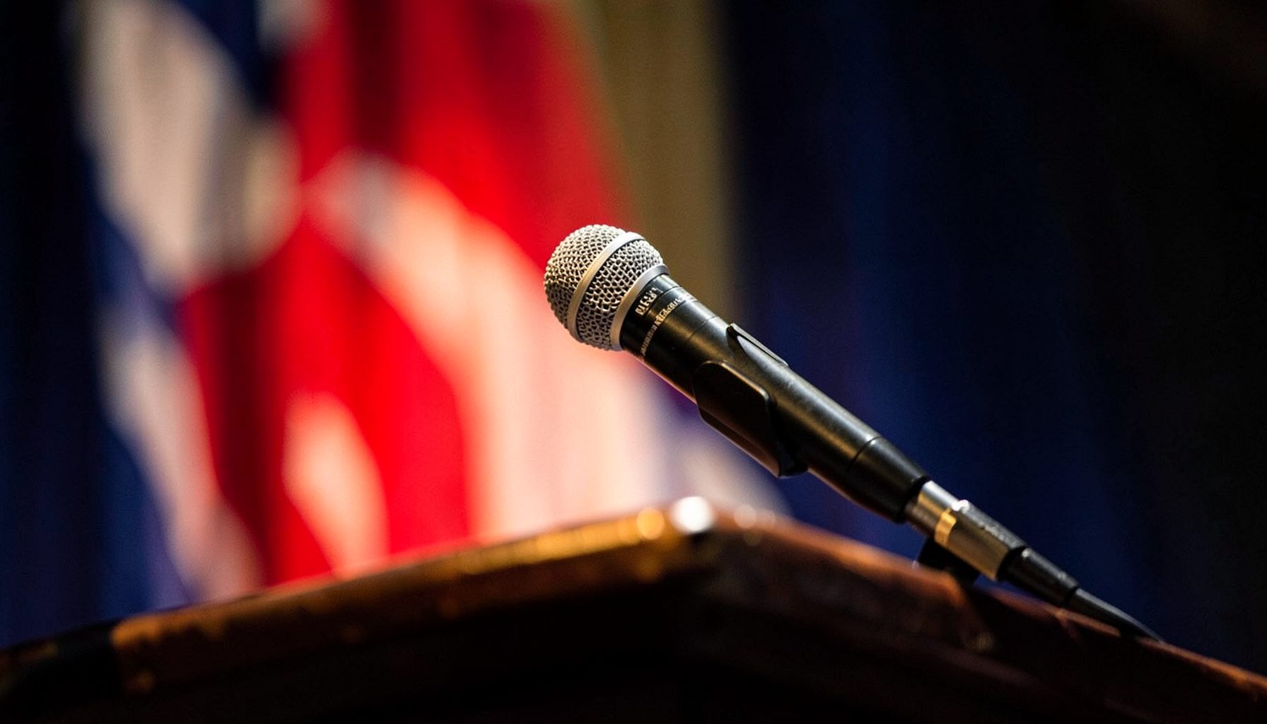 Close-up of a microphone on a stand atop a wooden podium, with a blurred flag in red, white, and blue in the background.
